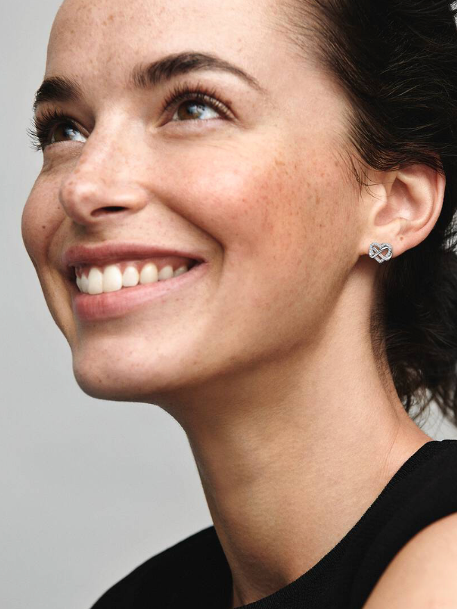 Close-up of a smiling woman wearing a small silver geometric dangle earring with clear stones, against a light gray background.