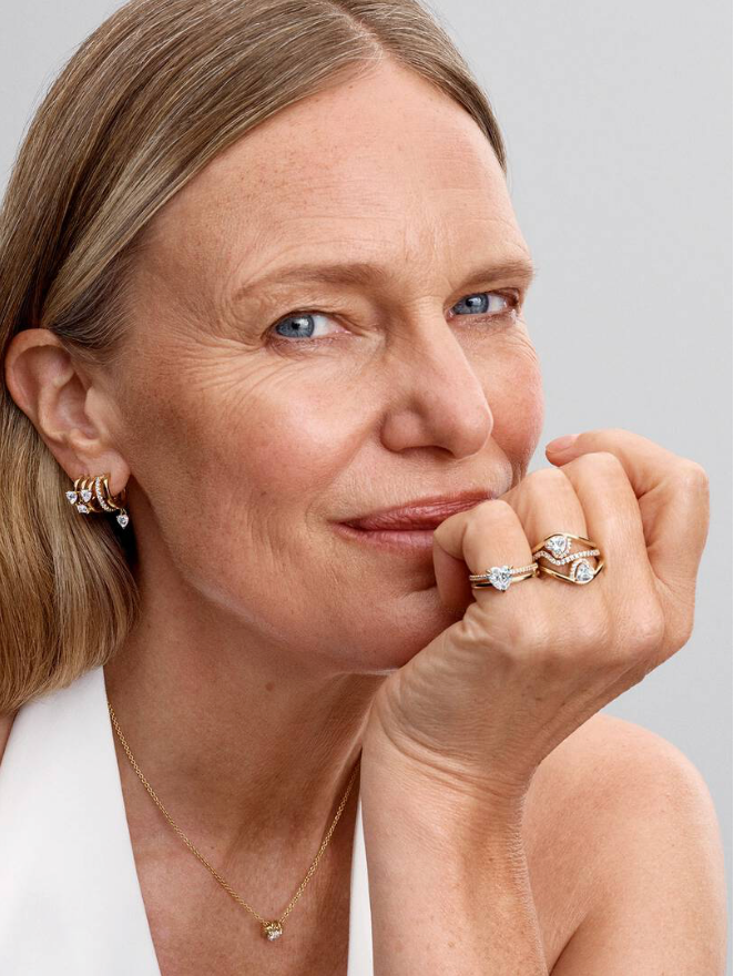 A close-up portrait of a woman wearing multiple pieces of jewelry, including a delicate gold necklace, a gold hoop earring with a clear gemstone, and two gold rings with clear gemstones on her fingers, set against a solid light gray background.