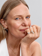 A close-up portrait of a woman wearing multiple pieces of jewelry, including a delicate gold necklace, a gold hoop earring with a clear gemstone, and two gold rings with clear gemstones on her fingers, set against a solid light gray background.