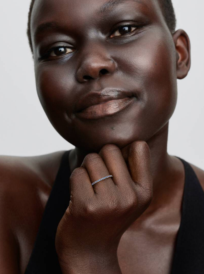 A close-up portrait of a model wearing a delicate silver band ring on her finger, set against a clean light gray background.