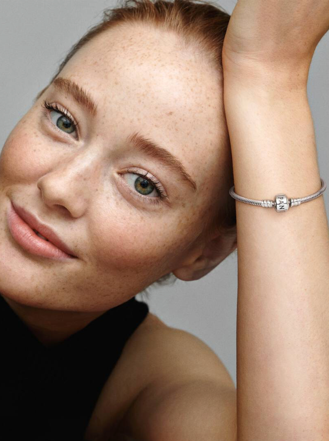 A close-up lifestyle shot of a young woman with freckles resting her head on her hand, wearing a delicate silver bracelet on her wrist against a neutral gray background.