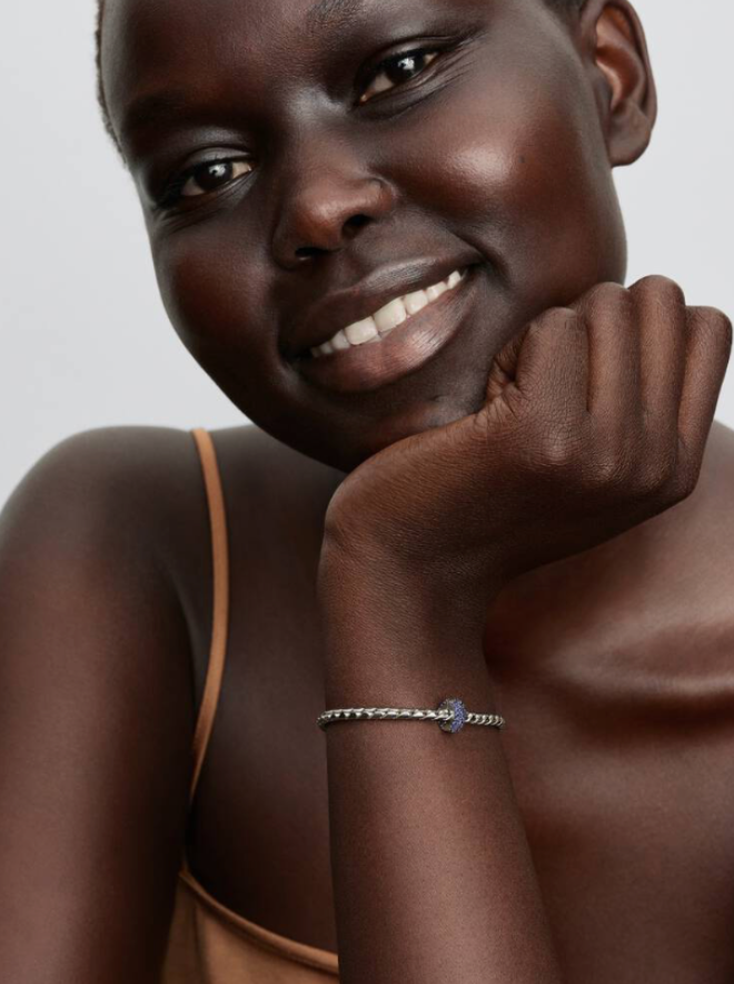 A close-up lifestyle shot of a smiling woman with dark skin resting her chin on her hand, wearing a delicate silver beaded bracelet with a small blue flower charm.