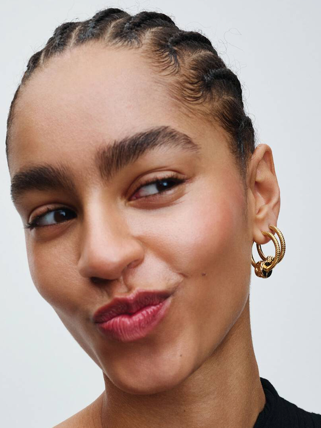 Close-up of a woman wearing a textured gold hoop earring, showing the side profile of her ear with the earring clearly visible against a light gray background.