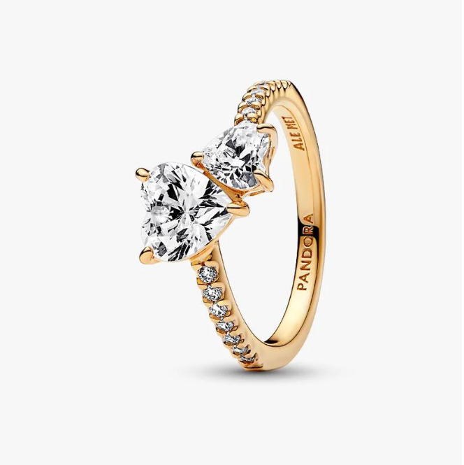 A gold ring with three heart-shaped clear gemstones and small accent stones along the band, shown on a clean white background. The brand name 'PANDORA' is engraved inside the band.