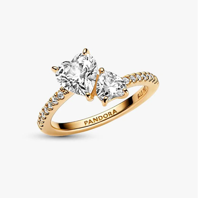A gold ring with two heart-shaped clear gemstones and small pavé accents on the band, shown on a light gray background. The inside of the band is engraved with the brand name PANDORA.
