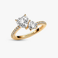 A gold ring with two heart-shaped clear gemstones and small pavé accents on the band, shown on a light gray background. The inside of the band is engraved with the brand name PANDORA.