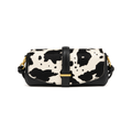 A small shoulder bag with a black and off-white cow print pattern on the front flap, solid black leather trim, a central black strap with a gold-tone buckle, and gold-tone side hardware, shown on a plain white background.